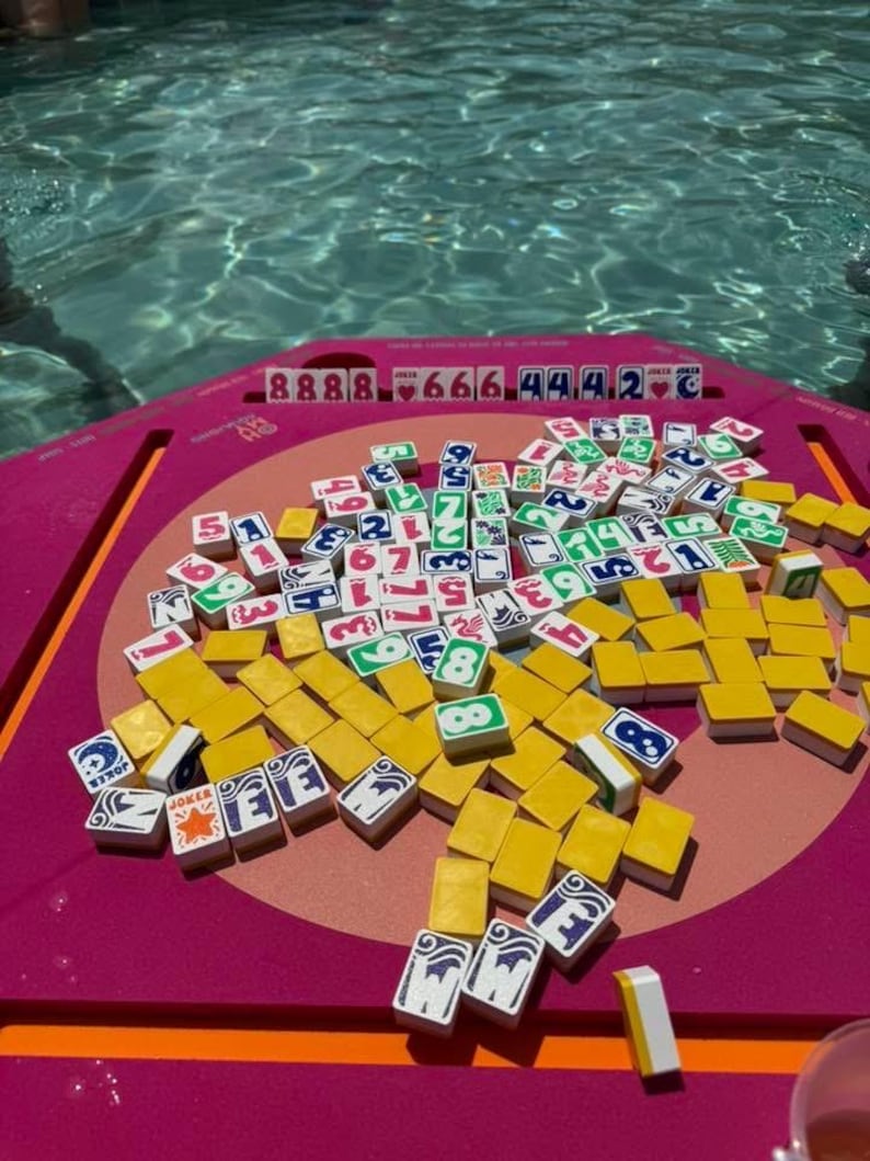 May include: A vibrant pink Mahjong game set with yellow tiles and various symbols and numbers. The tiles are scattered on a pink surface, with a background of a swimming pool. The game set includes tiles with the word "Joker".