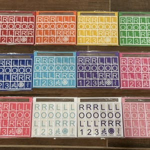 May include: A collection of colorful Scrabble tile sets, each in a different color, including red, orange, yellow, green, blue, teal, purple, pink, white, and light pink. Each set contains letter tiles and number tiles.