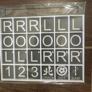 May include: A set of gray tiles with white letters, numbers, and symbols. The tiles spell out the letters R, L, and O, the numbers 1, 2, and 3, and the words CRACK, DOT, and BAM. The tiles are in a white plastic frame.
