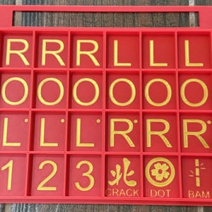 May include: A red game board with a handle, featuring gold letters, numbers, and symbols. The board is divided into squares, displaying letters like "R" and "L", numbers "1", "2", and "3", and symbols with the words "CRACK", "DOT", and "BAM".