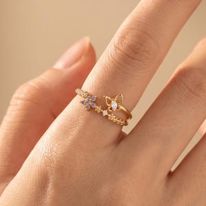 May include: A gold-toned open ring featuring a delicate butterfly motif with a central marquise-cut stone, alongside a small cluster of purple floral accents and tiny clear stones set along the thin, textured metal band.