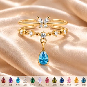 May include: A gold-toned ring featuring a double band design with a butterfly motif adorned with clear stones. The ring includes a dangling teardrop-shaped blue stone and a row of small clear stones. A chart below displays twelve birthstone options.