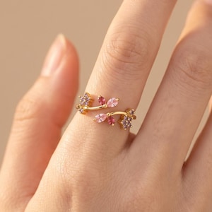 May include: A gold-toned open ring featuring a delicate vine-like design adorned with small, marquise-cut pink and purple gemstones. The band is thin and polished, with the stones arranged in clusters at each end of the open, overlapping structure.