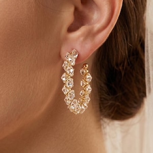 May include: Gold-toned hoop earrings with clear, faceted stones. The earrings are in a half-circle shape, with the stones closely set together. The earrings are worn on a person's ear, with a white veil in the background. The earrings are a bridal accessory.