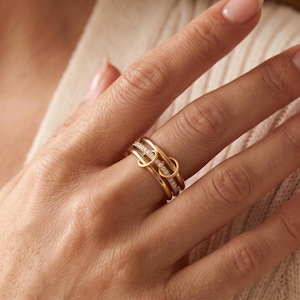 Peut inclure: Une bague en or à plusieurs anneaux avec une finition polie et une rangée centrale de petites pierres claires taillées en rond incrustées sur l'un des anneaux. Le design se compose de plusieurs boucles entrelacées et superposées.
