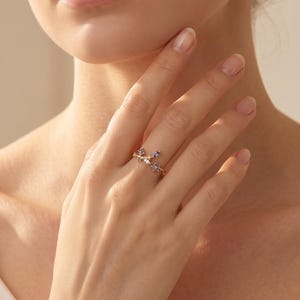 May include: A gold-toned ring featuring a delicate branch-like design adorned with small, multi-colored gemstones including blue, purple, and clear stones. The ring is worn on the ring finger, showcasing a slender band and a nature-inspired aesthetic.