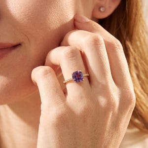 May include: A gold ring featuring a cushion-cut purple amethyst gemstone set in a four-prong setting. The band is thin and metallic, accented with small, clear pavé-set stones along the shank, creating a refined and classic jewelry piece.