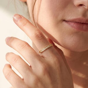 Peut inclure: Une fine bague en or ornée d'une rangée de petites pierres claires taillées en rond sur le dessus. Le métal présente une finition polie et réfléchissante, et les pierres sont disposées en ligne continue, offrant un aspect délicat et uniforme.