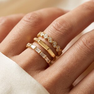 Peut inclure: Un ensemble de trois bagues en or portées ensemble. La bague supérieure présente un design ondulé avec de petites pierres rondes. La bague centrale est un anneau en or lisse et poli. La bague inférieure est sertie de pierres rectangulaires.
