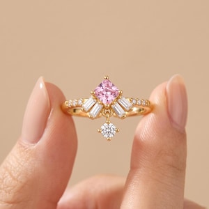Könnte beinhalten: Ein Goldring mit einem zentralen rosafarbenen Edelstein im Prinzessschliff, flankiert von weißen Baguette-Steinen auf beiden Seiten. Ein kleiner, runder, weißer Brillant hängt in der Mitte, und die Ringschiene ist mit winzigen weißen Steinen besetzt.