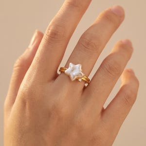 Könnte beinhalten: Ein goldfarbener Ring mit einer weißen, sternförmigen Perle als zentralem Blickfang. Die Ringschiene besteht aus zwei dünnen, parallelen goldfarbenen Drähten, die an der Basis des Sterns zusammenlaufen, der mit einem kleinen, klaren Stein verziert ist.
