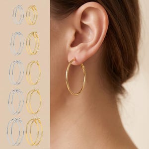 May include: A collection of thin, circular hoop earrings in silver and gold tones, displayed in multiple pairs on the left side, with a single gold hoop earring shown worn in an earlobe on the right side of the image.