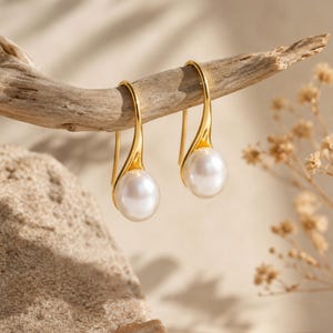 May include: A pair of gold-tone drop earrings featuring smooth, round white pearls suspended from curved metal hooks. The polished gold metal forms a sleek, elongated teardrop shape that cradles the base of each pearl, creating a refined look.