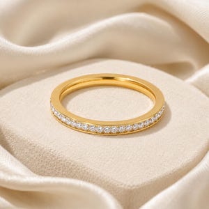 Könnte beinhalten: Ein goldfarbener Ring mit einem Band aus kleinen, klaren Edelsteinen. Der Ring ist vor einem neutralen Hintergrund platziert, der seine polierte Oberfläche und das Funkeln der Steine hervorhebt. Der Ring ist ein zartes Schmuckstück.