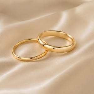 May include: Two polished gold rings with smooth, rounded bands. One ring features a slightly thicker profile than the other. Both bands have a high-shine, reflective finish that catches the light, showcasing a simple, continuous circular design.