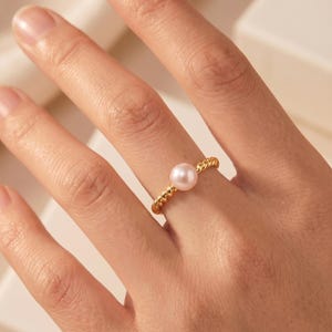 Puede incluir: Un anillo de oro con una banda trenzada estilo cuerda y una perla única, lisa, redonda y de color rosa pálido centrada en la parte superior. El metal tiene un acabado de oro amarillo pulido y la perla muestra una textura suave y brillante.