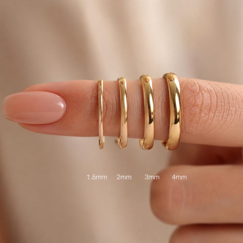 May include: Four gold-toned rings with smooth, polished surfaces, displayed in a row with widths labeled as 1.5mm, 2mm, 3mm, and 4mm. The rings are arranged by increasing thickness, showcasing a simple, rounded band design with a reflective finish.