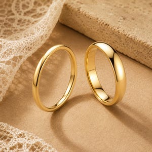 May include: Two polished gold rings with a smooth, rounded surface. The rings feature a simple, solid band design with a high-shine metallic finish that reflects light, creating bright highlights along the curved edges of the circular metal bands.