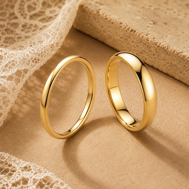 May include: Two polished gold rings with a smooth, rounded surface. The rings feature a simple, solid band design with a high-shine metallic finish that reflects light, creating bright highlights along the curved edges of the circular metal bands.