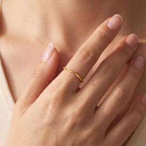 May include: A thin, polished gold ring featuring a continuous twisted or faceted texture along the band. The ring is worn on the middle finger, showcasing a simple, slender design with a reflective metallic finish that catches the light.