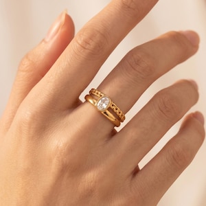 May include: A gold-toned double band ring featuring a smooth, plain band paired with a textured chain-link band. The ring is centered with a single, clear, oval-shaped gemstone set in a polished gold-toned bezel, creating a layered appearance.