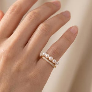 May include: Two gold rings are stacked on a finger. The top ring features a row of white pearls, while the bottom ring is adorned with small, sparkling stones. The rings are set against a neutral background.