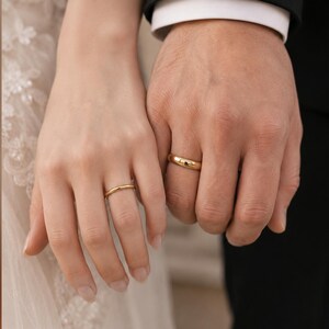 May include: Two gold wedding bands with a smooth, polished finish. One ring is thinner and worn on the left hand, while the other is wider and worn on the right hand. Both rings feature a simple, rounded profile and a bright, reflective yellow gold surface.