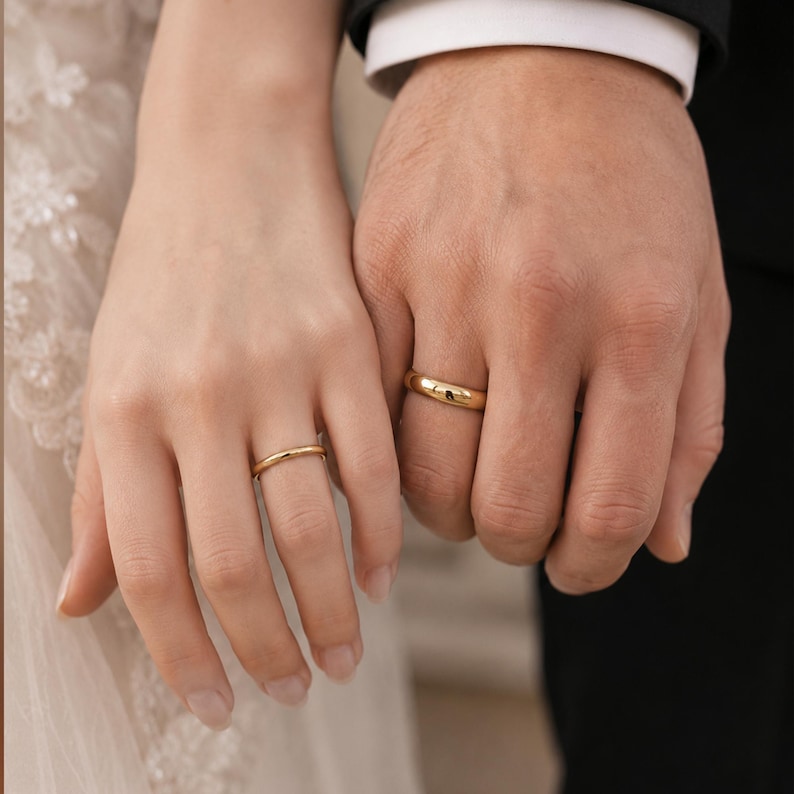 May include: Two gold wedding bands with a smooth, polished finish. One ring is thinner and worn on the left hand, while the other is wider and worn on the right hand. Both rings feature a simple, rounded profile and a bright, reflective yellow gold surface.