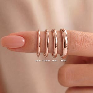 以下が含まれることがあります： 滑らかで光沢のある表面を持つ4つのローズゴールドのリングが、厚さの順に並んでいます。各リングには1mm、1.5mm、2mm、3mmの幅がラベル付けされており、バンドサイズの明確な視覚的比較を提供しています。