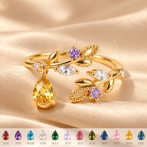 Puede incluir: Un anillo abierto de tono dorado con dos ramas en forma de hoja adornadas con pequeñas piedras redondas y de talla marquesa transparentes y moradas. Una piedra amarilla en forma de pera cuelga de una rama. Una tabla muestra doce piedras de nacimiento.