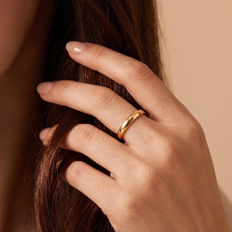 May include: A polished gold band ring with a smooth, rounded surface and a high-shine finish. The ring features a simple, solid construction with a consistent width throughout the entire circumference, reflecting light across its curved metallic surface.