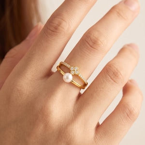 Könnte beinhalten: Ein goldener Doppelring mit einer einzelnen weißen Perle und einem vierblättrigen Blumenmotiv, das mit kleinen klaren Steinen besetzt ist. Das obere Band ist mit winzigen klaren Steinen verziert, während das untere Band die Perle in einer Fassung hält.