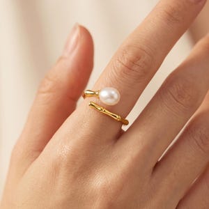 May include: A gold-toned open ring featuring a textured bamboo-inspired band and a single white oval-shaped pearl at one end. The ring is designed with a bypass style, allowing the two ends to overlap slightly while remaining open at the finger.