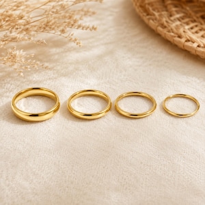 May include: A set of four polished gold-tone metal rings arranged in a horizontal row, ranging in thickness from the widest ring on the left to the thinnest ring on the right, each featuring a smooth, rounded band and a reflective, metallic finish.