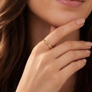 May include: A simple, polished gold band ring with a smooth, rounded surface. The ring features a continuous, solid metal construction with a high-shine finish, worn on a finger to showcase its slim, circular profile and reflective yellow gold tone.