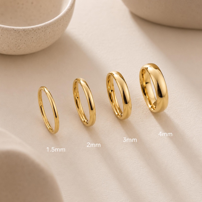 May include: Four polished gold-tone rings arranged in a row, labeled with widths of 1.5mm, 2mm, 3mm, and 4mm. Each ring features a smooth, rounded band design with a reflective metallic finish, showcasing the varying thickness of the jewelry pieces.