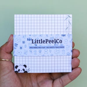 May include: A square memo pad with a white grid pattern, featuring a small panda illustration in the bottom left corner. A blue paper band wraps around the center, printed with the text LittlePeelCo, MINI MEMO PAD 4x4, 50 SHEETS, 24LB PAPER.