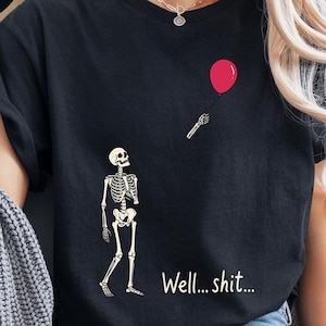 May include: A black t-shirt featuring a white skeleton graphic looking up at a detached skeletal hand holding a red balloon. The text "Well... shit..." is printed in white below the skeleton. The shirt has short sleeves and a crew neckline.