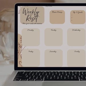 Può includere: Un computer portatile mostra un pianificatore settimanale digitale. Il pianificatore ha il testo "Weekly Reset" e include sezioni per ogni giorno, un'area "Brain Dump" e "Top 3 Goals". È visibile anche il testo "Pianifica la tua settimana con intenzione".