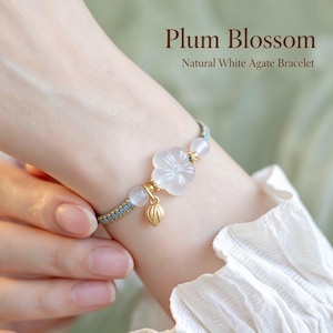 May include: A bracelet featuring a carved white agate plum blossom, two round white agate beads, and a small gold-toned metal leaf charm on a blue braided cord. The text Plum Blossom Natural White Agate Bracelet is visible.