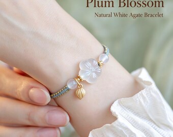 Puede incluir: Una pulsera con una flor de ciruelo de ágata blanca tallada, dos cuentas redondas de ágata blanca y un pequeño dije de hoja de metal dorado en un cordón trenzado azul. El texto Plum Blossom Natural White Agate Bracelet es visible.