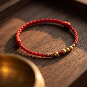 May include: A red braided cord bracelet featuring three smooth, matte gold-toned spherical beads. The bracelet includes small sections of multi-colored thread wrapping near the beads and adjustable knotted ends for a secure fit.