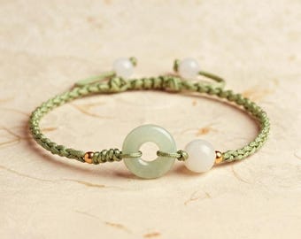 Puede incluir: Una pulsera con un cordón trenzado de color verde salvia, un anillo central de color verde pálido similar al jade, una cuenta blanca y pequeños detalles metálicos dorados. El cordón ajustable termina en dos cuentas blancas, creando una joya sencilla.