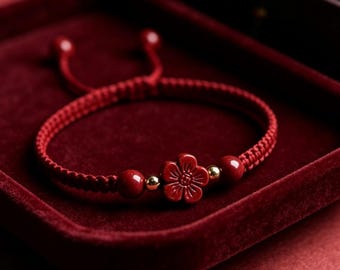Puede incluir: Una pulsera de cordón trenzado rojo con una cuenta central tallada en forma de flor roja, flanqueada por dos cuentas esféricas rojas más pequeñas y dos diminutas cuentas metálicas doradas. La pulsera tiene un cierre de nudo deslizante.