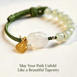 Puede incluir: Una pulsera con un cordón trenzado verde, una serie de cuentas de color verde pálido, un dije de conejo blanco tallado y un pequeño colgante redondo de tono dorado con caracteres chinos. El texto dice: May Your Path Unfold Like a Beautiful Tapestry.