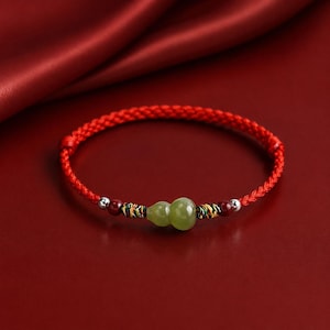 May include: A red braided cord bracelet featuring a central light green gourd-shaped stone bead. The design includes small dark red spherical beads, metallic silver-toned accents, and decorative multi-colored thread wraps flanking the gourd.