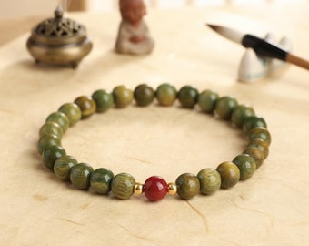 Puede incluir: Una pulsera de cuentas con cuentas redondas de madera verde, una cuenta roja central única y dos pequeños espaciadores de metal dorado. Las cuentas están ensartadas en un cordón elástico, creando una forma circular con acabado pulido.