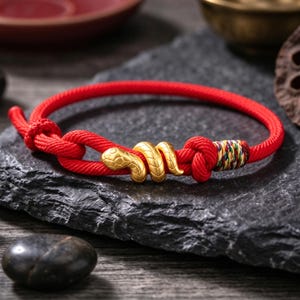 May include: A red cord bracelet featuring a central gold-coloured snake charm. The bracelet is secured with two knots and includes a small section of multicoloured thread wrapping near one of the knots, creating a distinct visual accent.