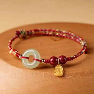 May include: A red and gold bracelet featuring a pale green circular jade pendant, two red beads, and a small gold charm. The bracelet is made of braided red and gold threads.