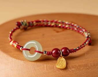Puede incluir: Una pulsera roja y dorada con un colgante de jade circular verde pálido, dos cuentas rojas y un pequeño dije dorado. La pulsera está hecha de hilos trenzados rojos y dorados.